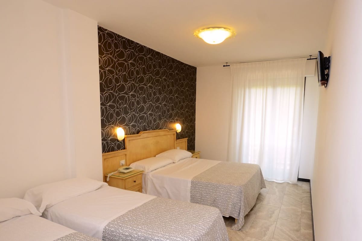 A hotel room with three beds, a patterned wall, a ceiling light, and a window with curtains.