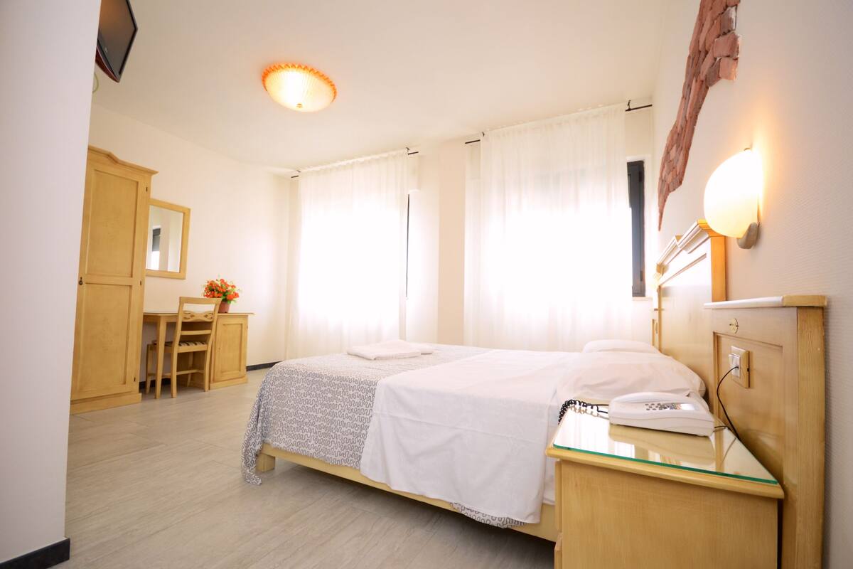 A hotel room with a bed, bedside table, lamp, and a desk with a chair.