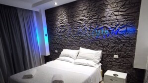 A modern bedroom with a stone-textured headboard, a neatly made bed with white linens, and a small bedside table with a lamp. - Hotel Clinton (Casoria)