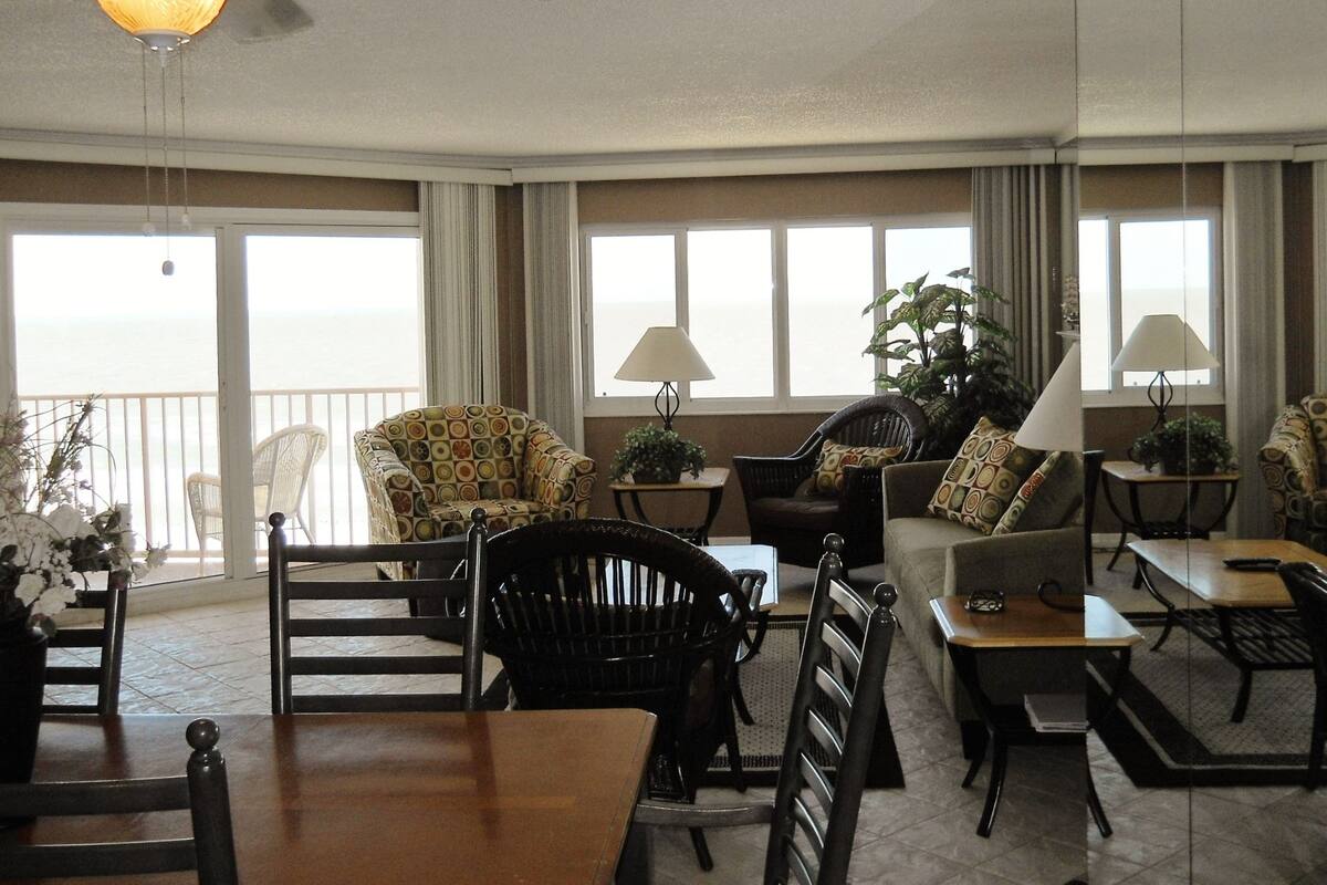 A spacious living room with a balcony overlooking the sea, furnished with various chairs and tables, and a ceiling fan.