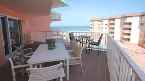 Un balcón con mesa y sillas, vista a la playa y un edificio al fondo.
