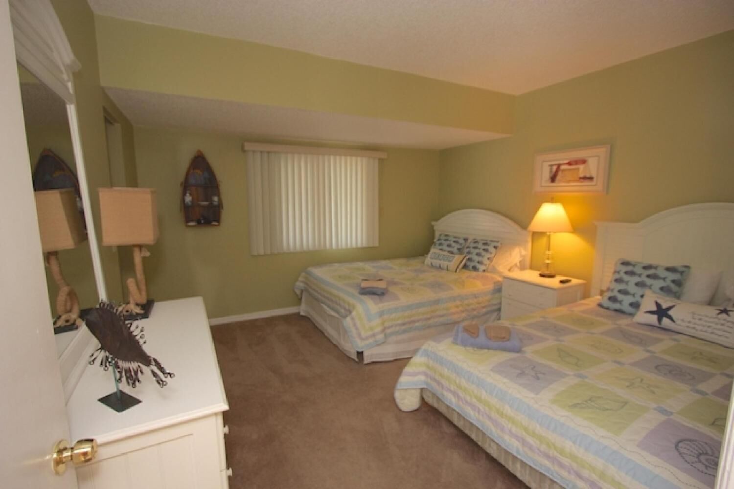 A bedroom with two beds, a dresser, a lamp, and a window with blinds.