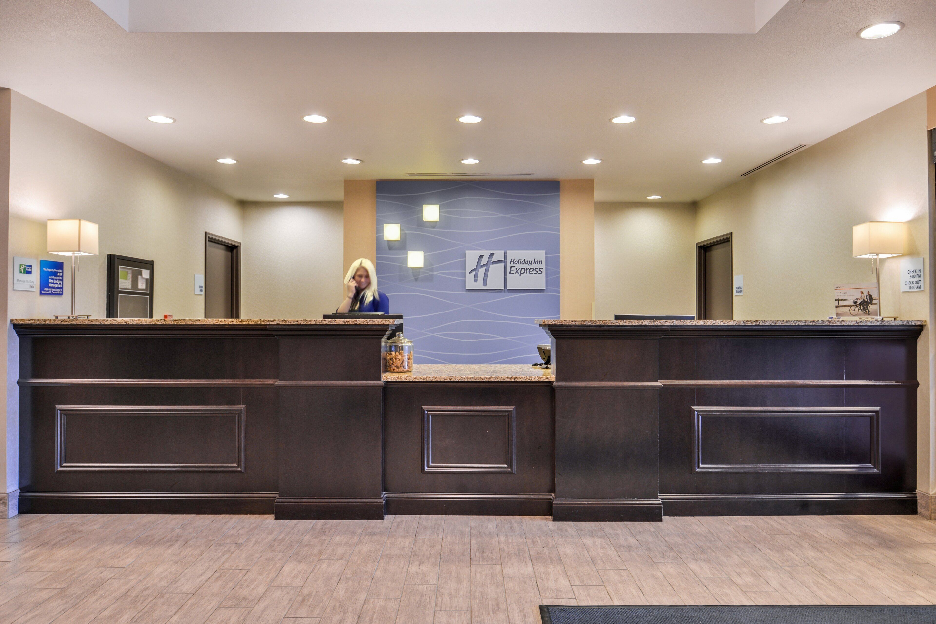 Una recepción de hotel con una recepcionista, un letrero de Holiday Inn Express y un horario de registro de entrada y salida.
