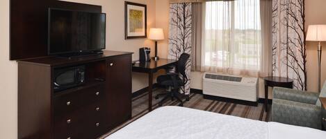 A hotel room with a bed, a desk with a chair, a television, a microwave, a lamp, and a window with curtains.