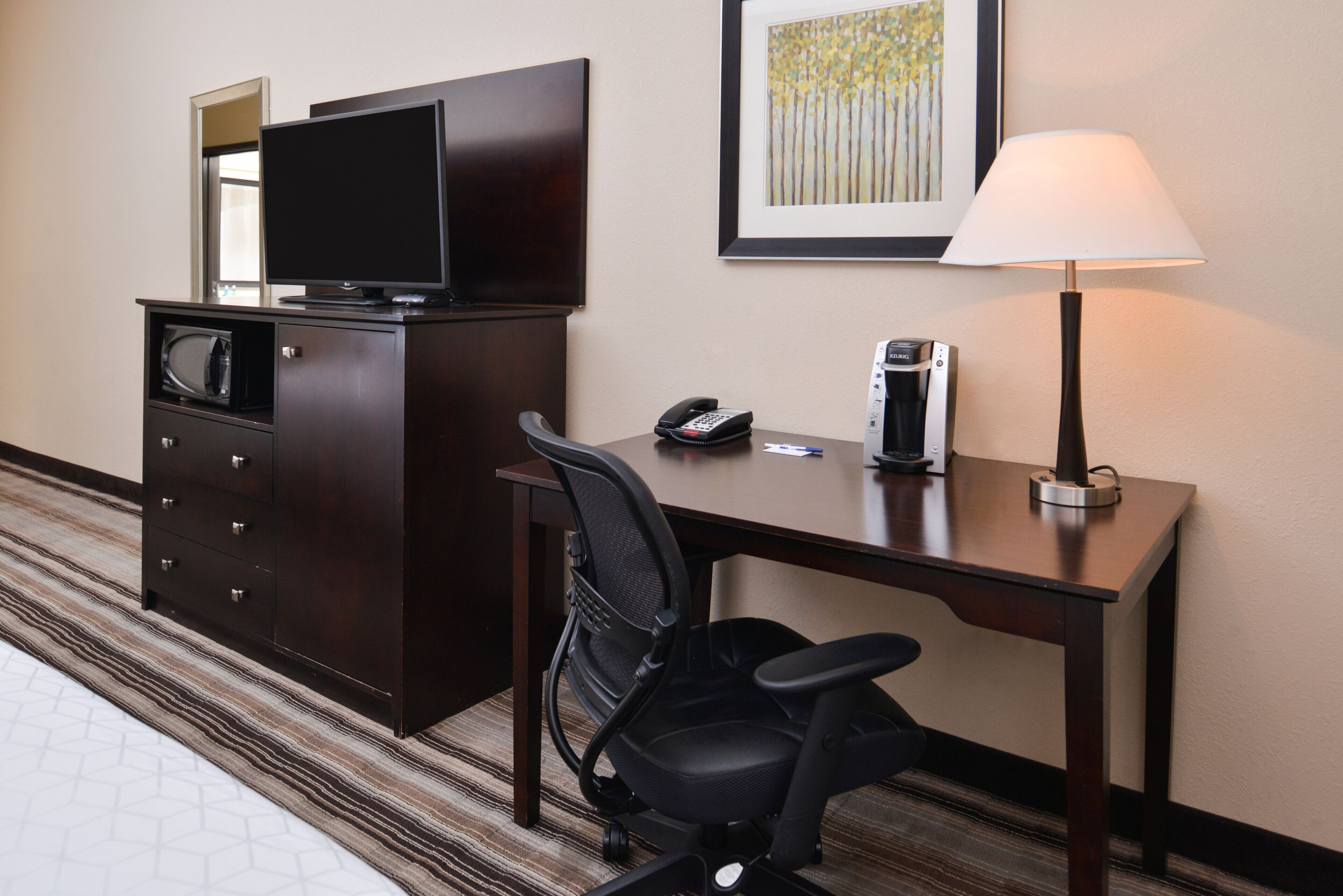 A hotel room with a desk, a chair, a television, a coffee maker, and a lamp.