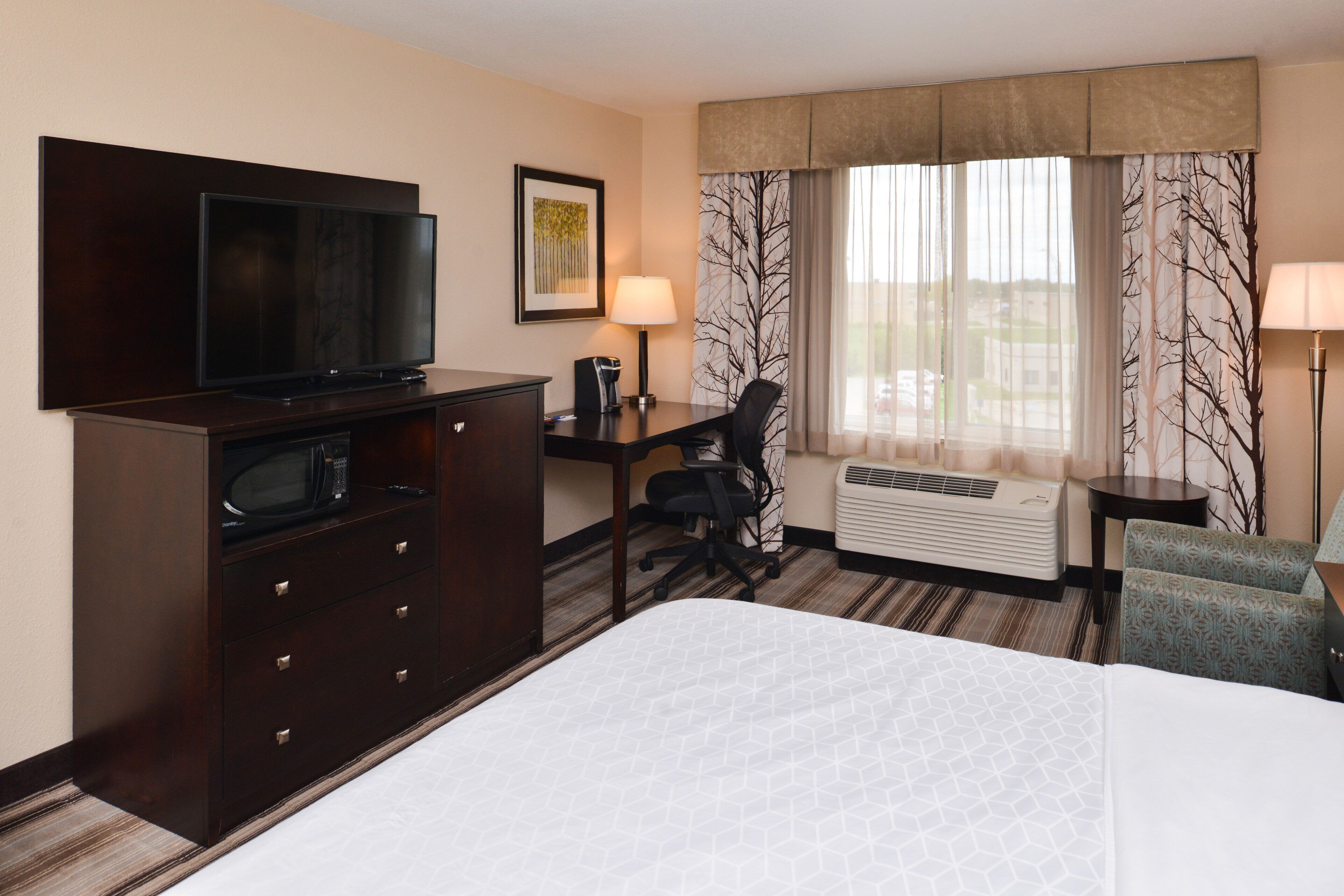 Habitación de hotel con cama, escritorio con silla, televisor, microondas, lámpara y ventana con cortinas.
