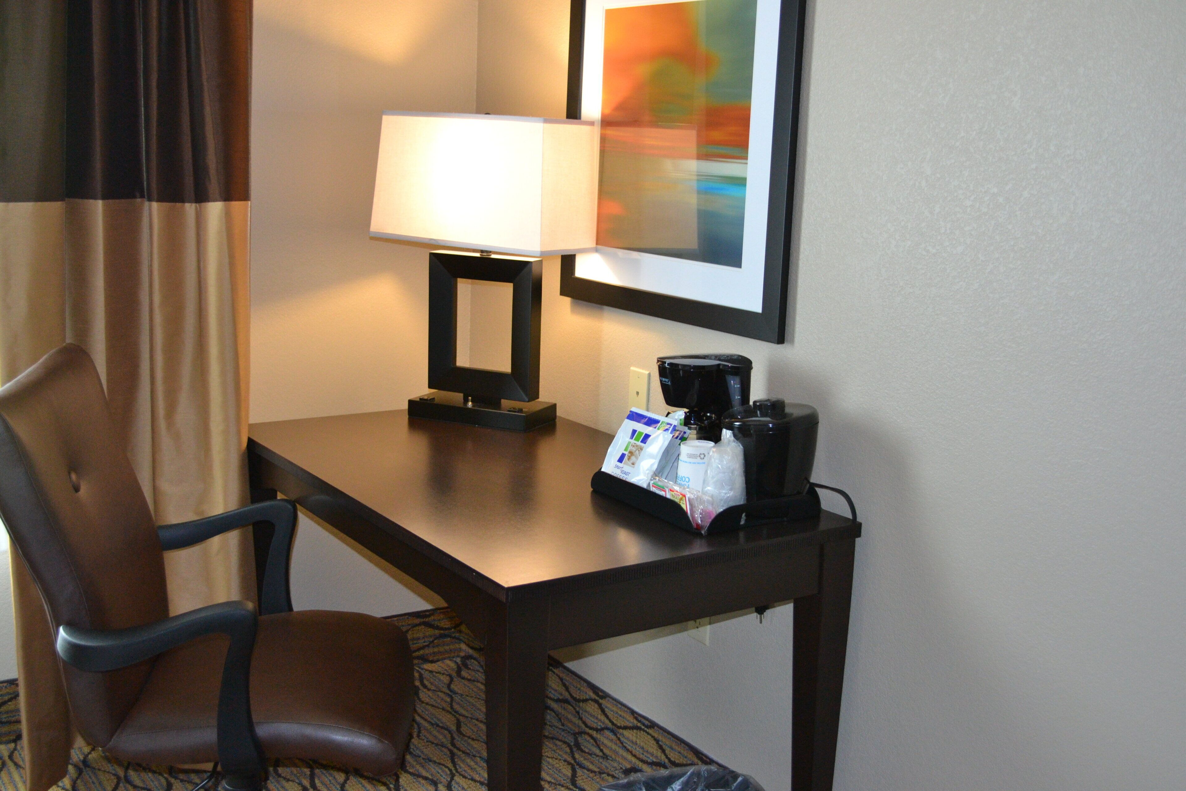 Habitación de hotel con escritorio, silla, cafetera y un cuadro en la pared.