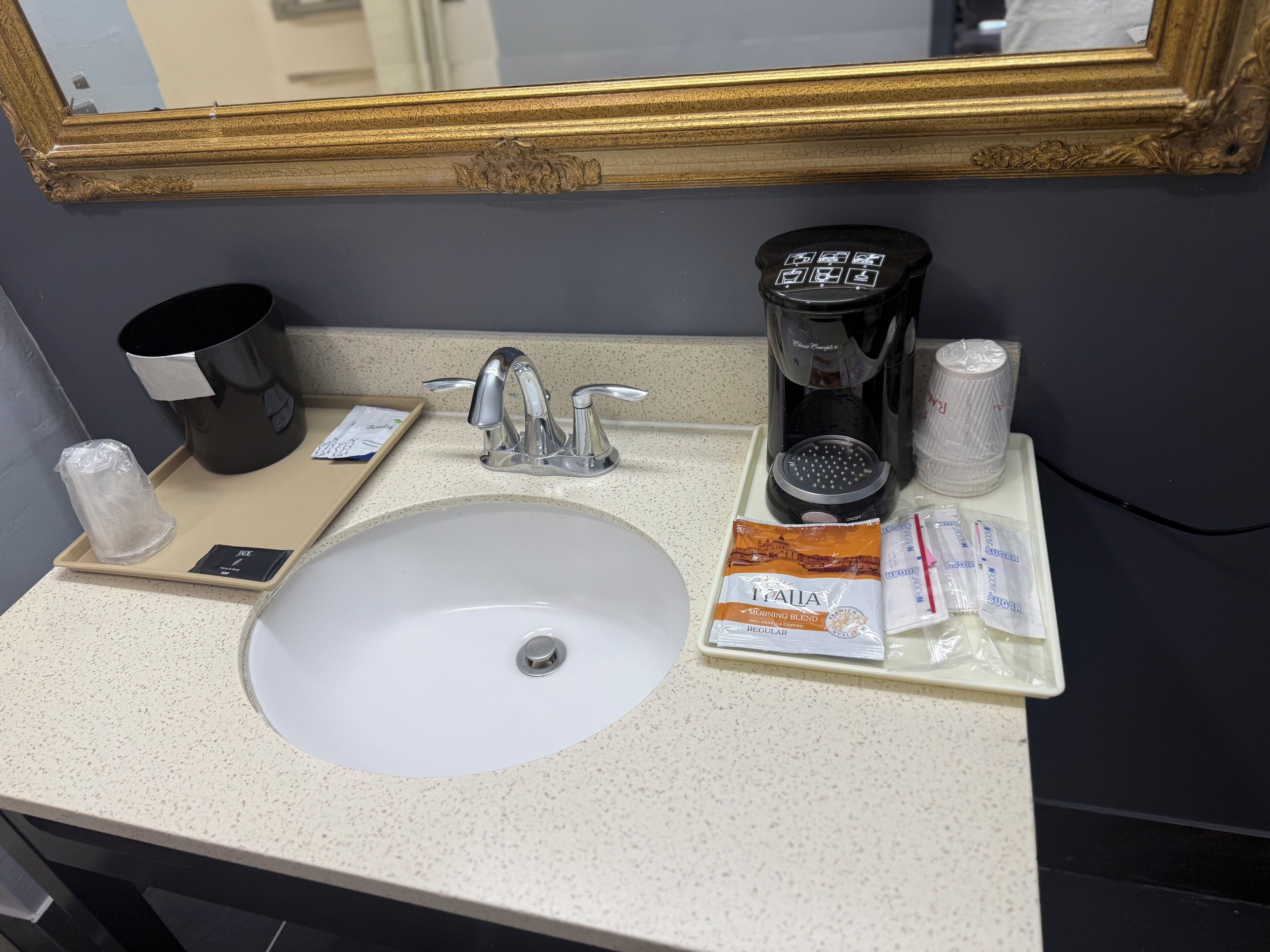 A hotel bathroom sink with a coffee maker, toiletries, and a trash bin.
