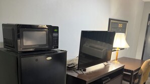 A compact hotel room with a kitchenette area featuring a refrigerator, microwave, and a small TV on top. There is a desk with a lamp and a chair, and a bed with a patterned bedspread. - Western Motel (Jesup)