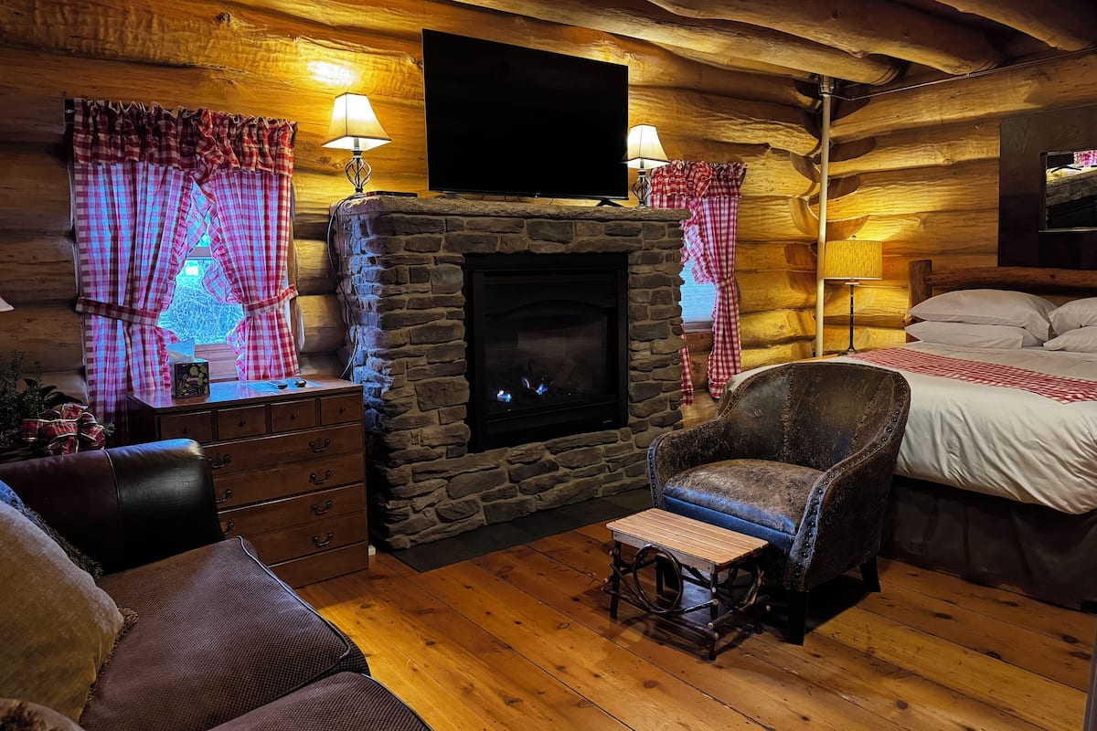 A cozy log cabin room with a stone fireplace, a comfortable sofa, a wooden coffee table, and a bed with checkered curtains.