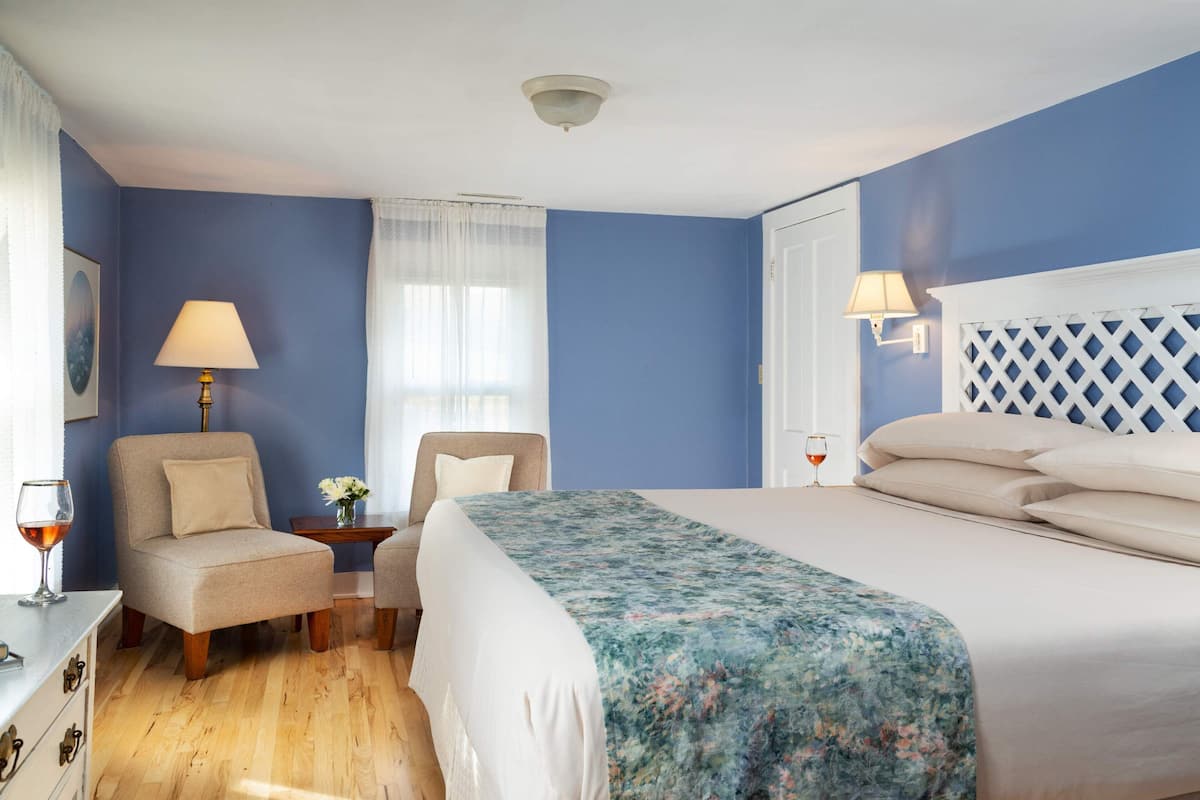 A hotel room with a bed, two chairs, a small table with a lamp, and a window with curtains.