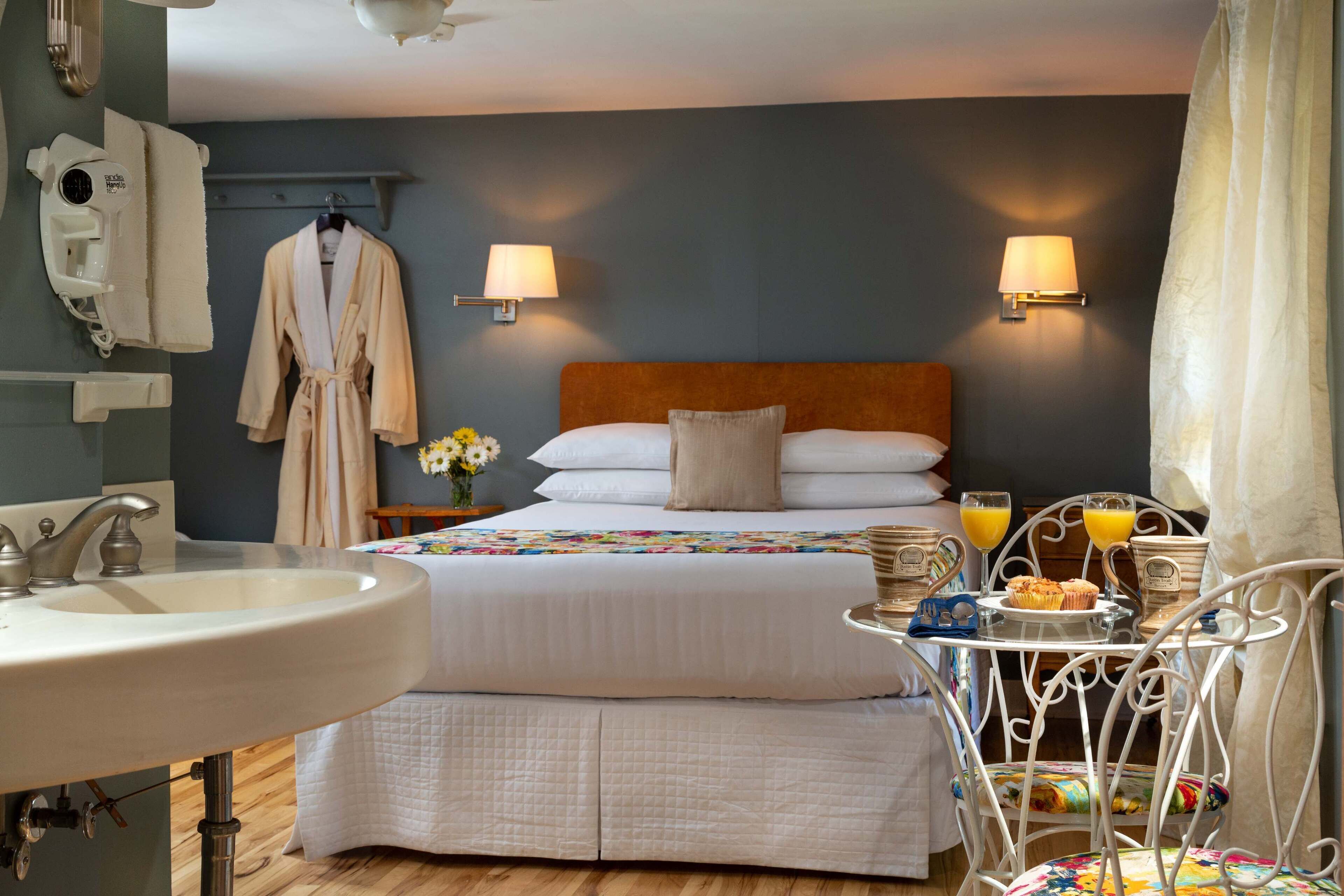 Habitación de hotel con cama, lámparas de noche, lavamanos y una mesa de desayuno con muffins y jugo.