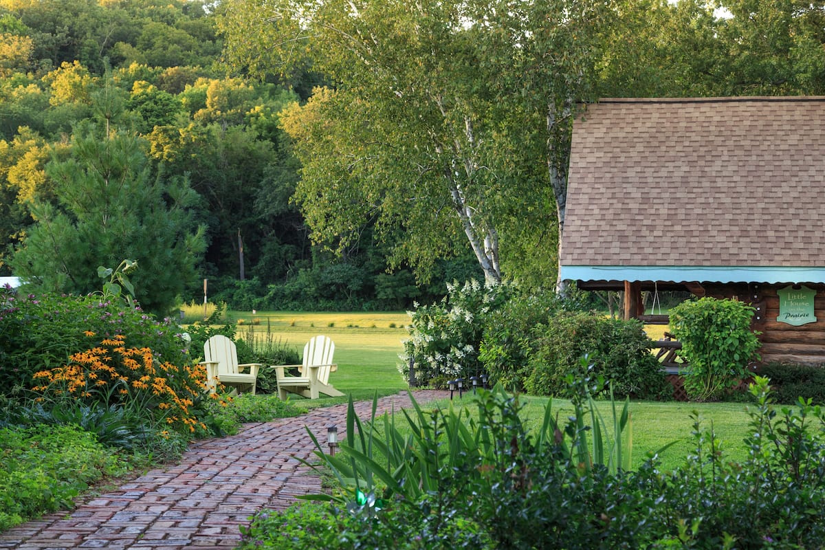 A rustic cabin with a brick pathway leading to a wooden deck, surrounded by lush greenery and a well-maintained lawn.