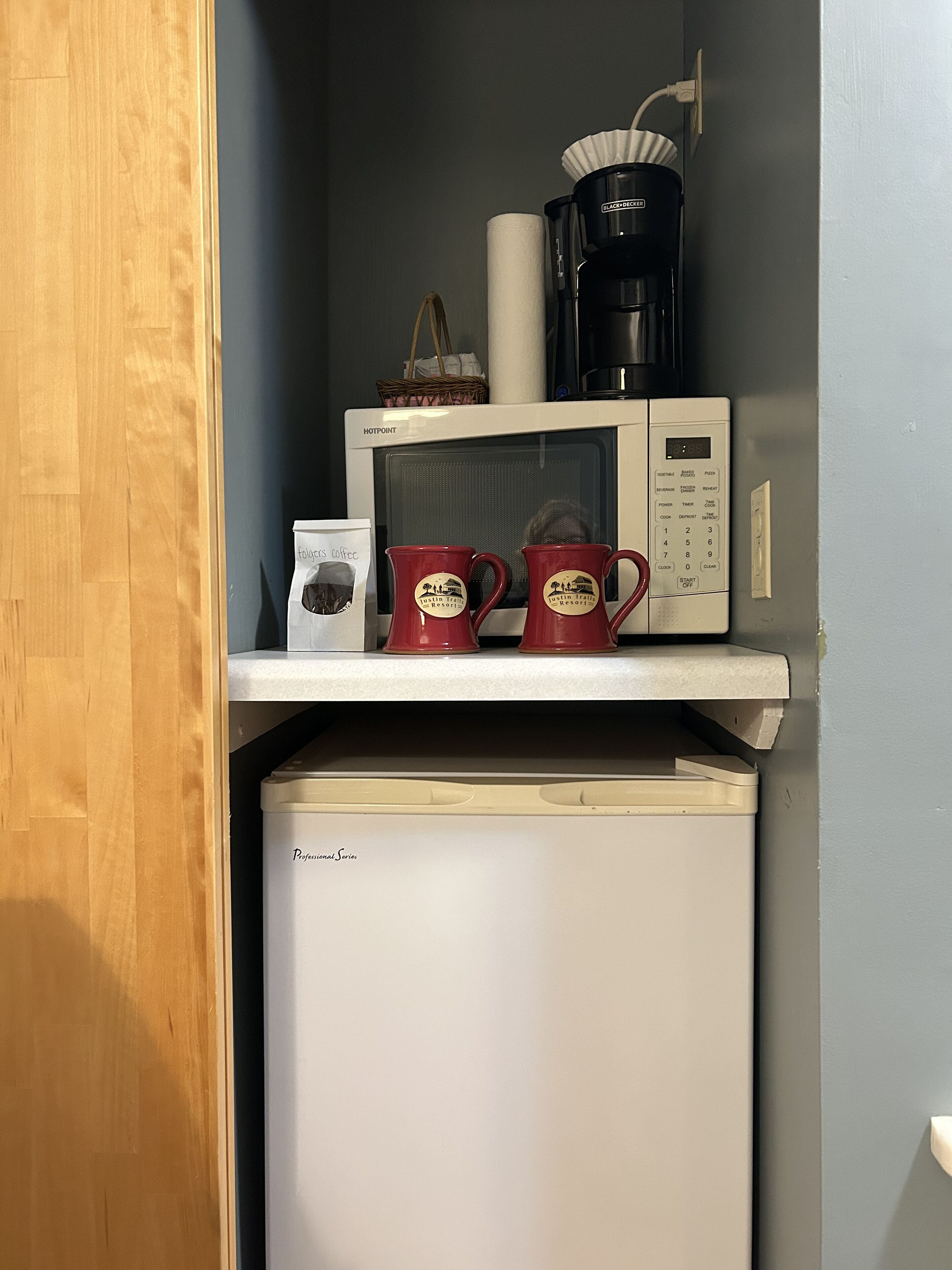 Uma cozinha compacta com micro-ondas, cafeteira e duas canecas vermelhas em uma prateleira.