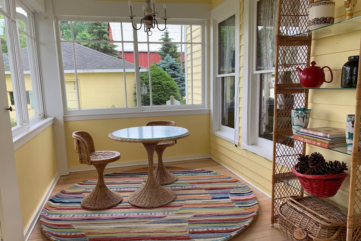 A cozy room with a round table and wicker chairs, a colorful striped rug, and a wicker shelf with books and decorative items.