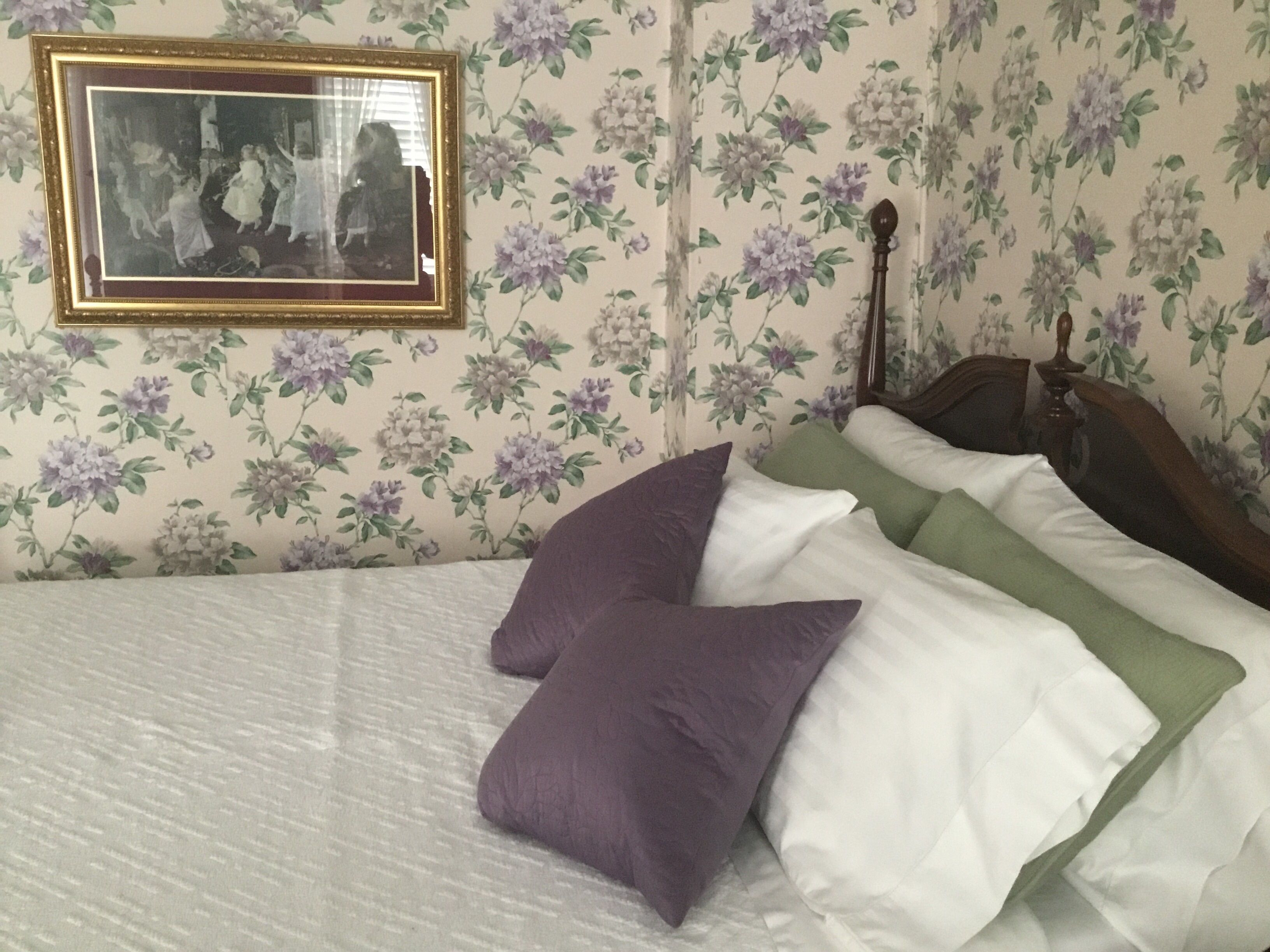 Quarto com papel de parede floral, cama com roupa de cama branca e verde, e uma moldura com imagem na parede.