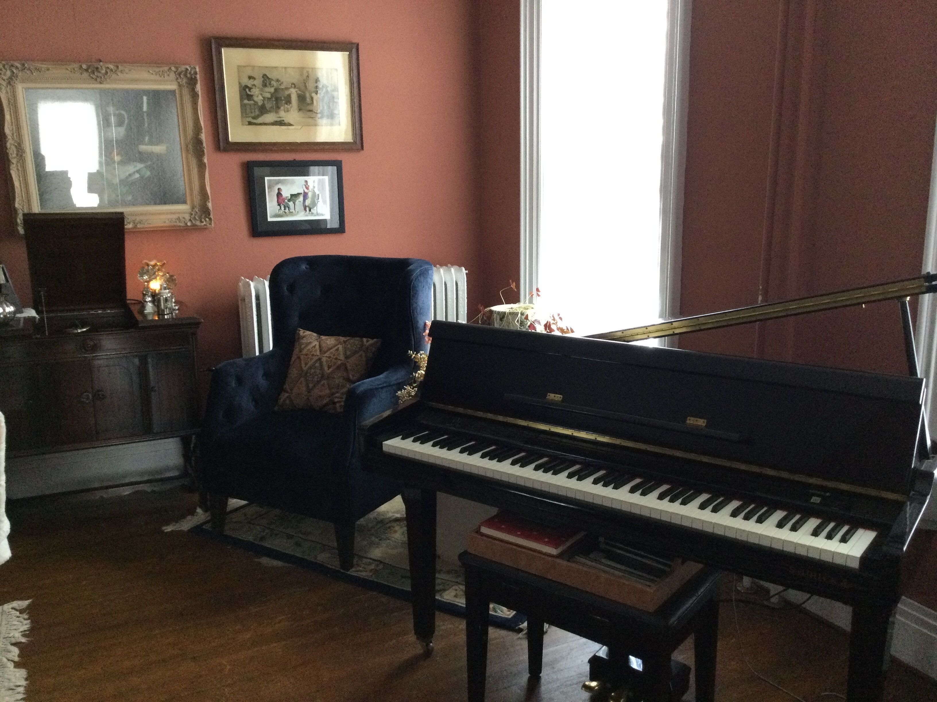 A room with a grand piano, a dark blue armchair, a radiator, and framed pictures on the wall.