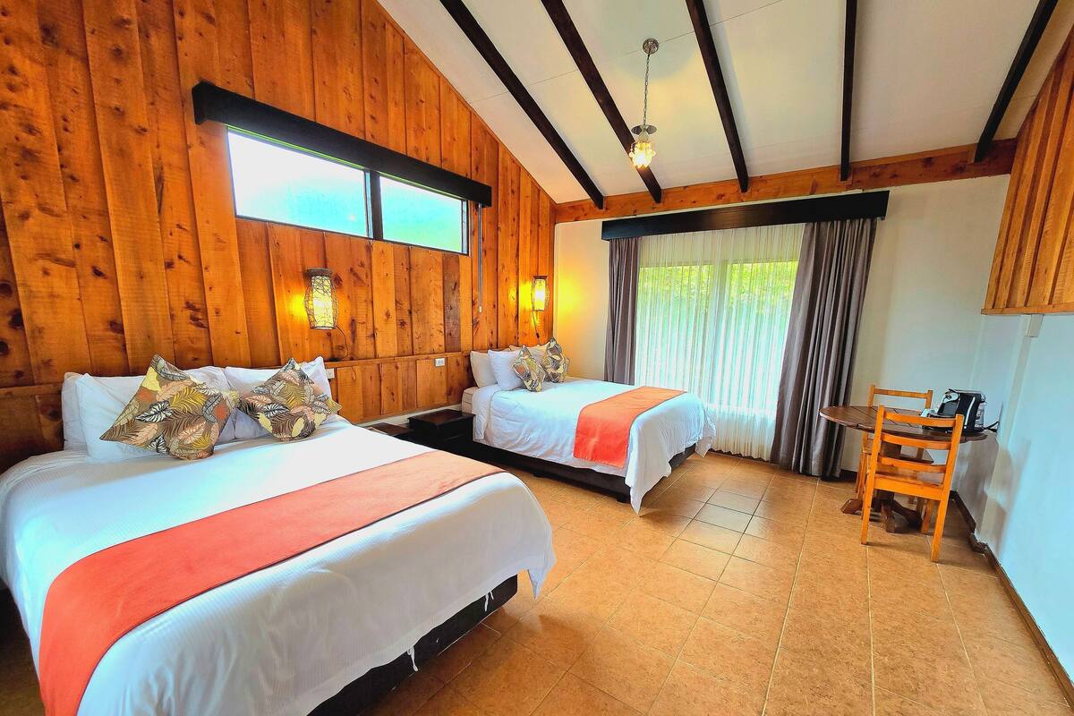 Una habitación con dos camas, paredes de madera, una mesa de comedor con sillas y un ventanal con cortinas.
