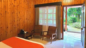 Een houten kamer met een bed, twee stoelen, een tafel met een koffiezetapparaat, en een deur die uitkomt op een tuin.