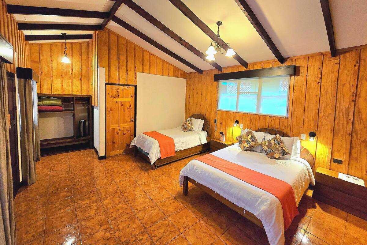 Una habitación acogedora con paredes de madera, una cama grande, dos camas más pequeñas, un ventanal con vistas y un ventilador de techo.