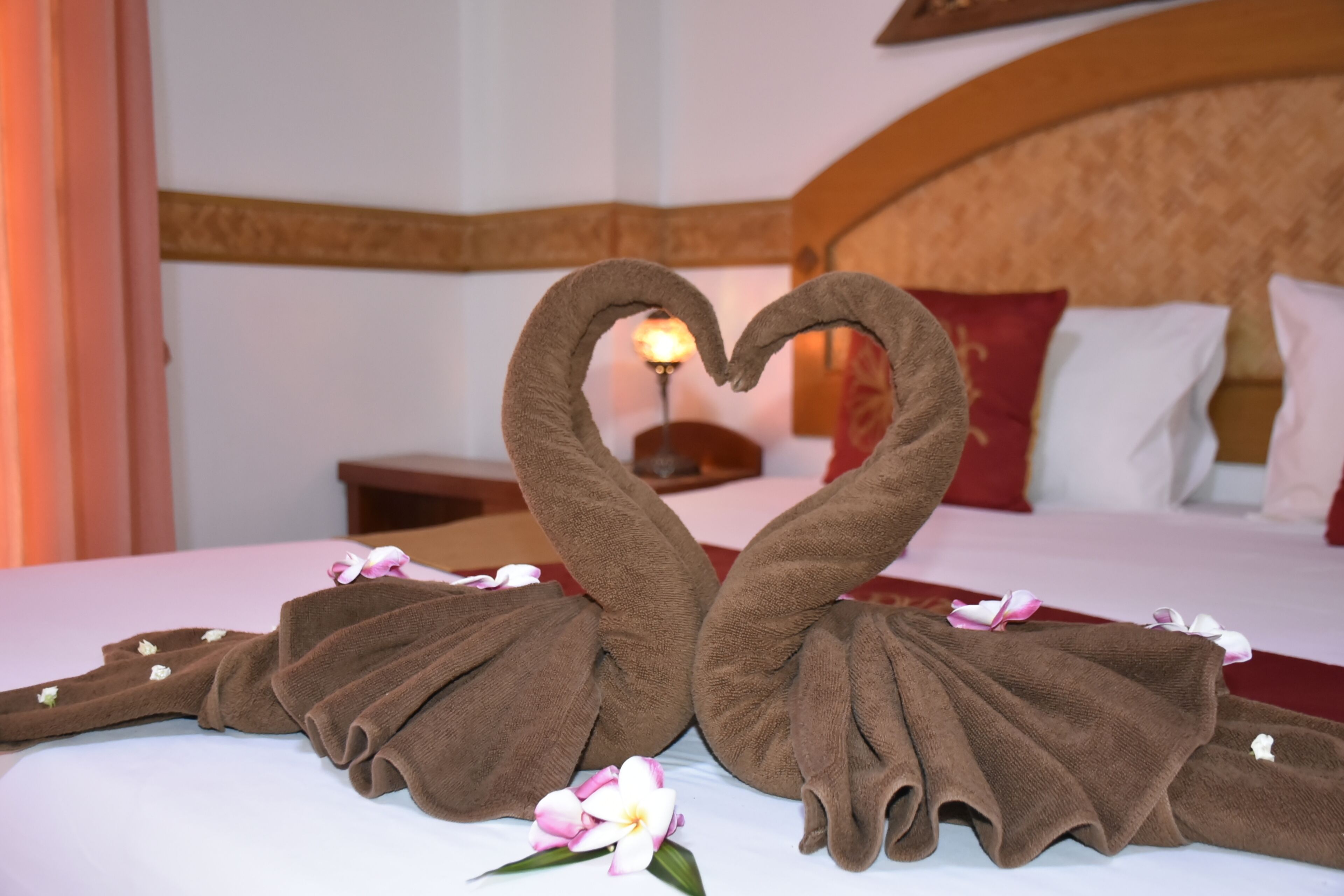 Ein Bett mit einer Handtuch-Arrangement in Herzform, einer Blume und einer Lampe im Hintergrund.