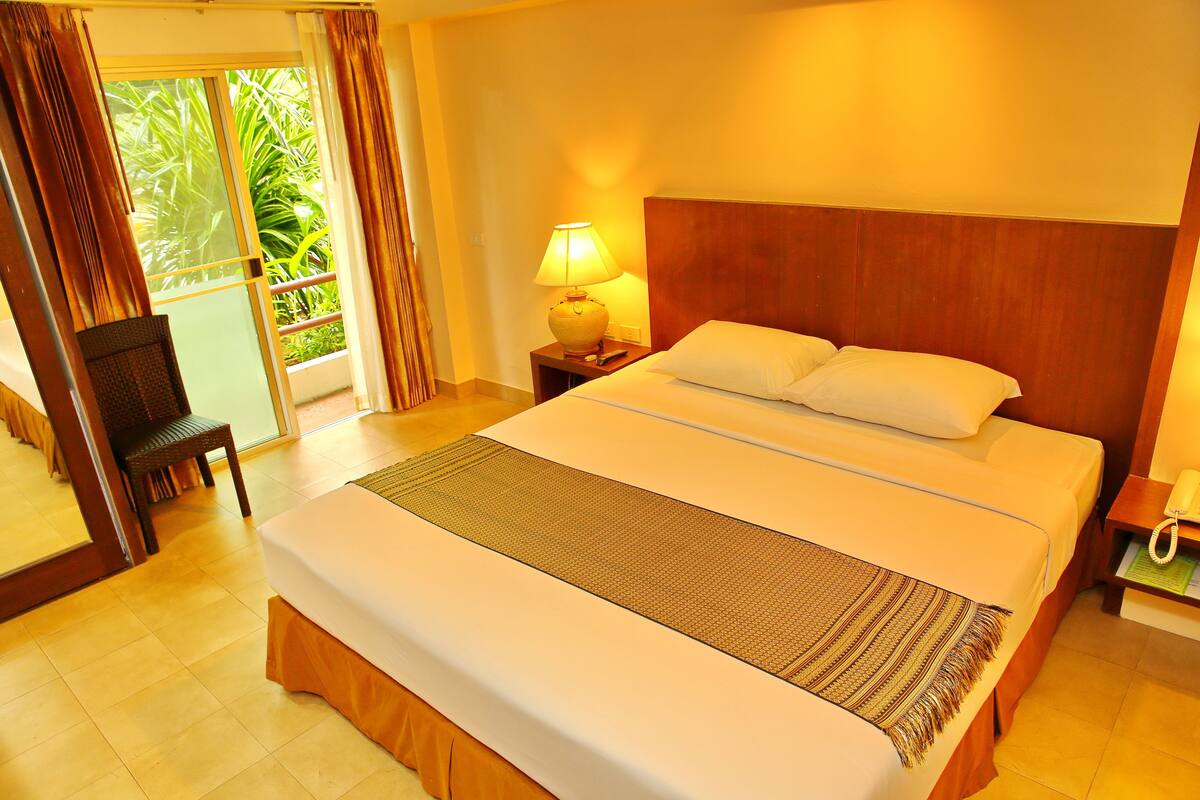 Habitación de hotel con una cama grande, una mesita de noche con lámpara, una silla y vista a zonas verdes por la ventana.