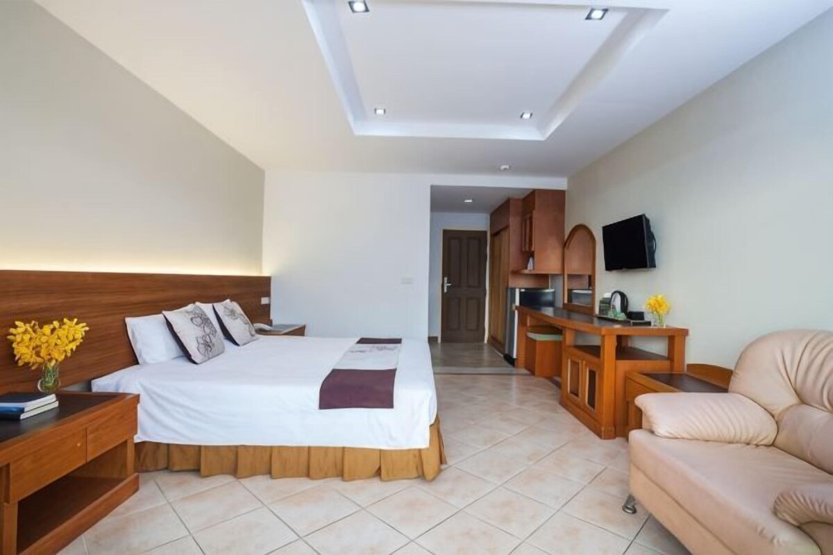 Une chambre d’hôtel avec un lit, une table de chevet en bois, un canapé beige, une petite table avec un vase et un téléviseur fixé au mur.