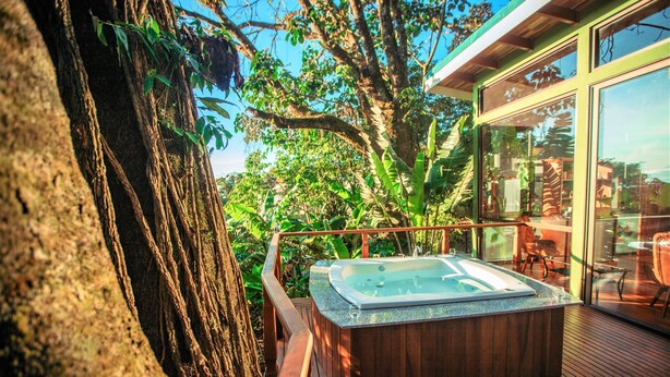 Un jacuzzi en una terraza de madera con barandilla de vidrio, con vistas a un jardín exuberante y un edificio moderno.