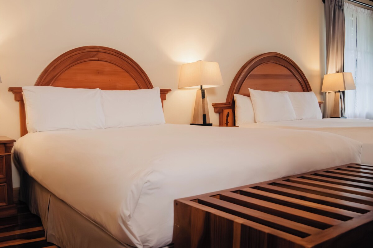 A hotel room with two beds, wooden headboards, bedside lamps, a wooden bench, and a nightstand with a drawer.