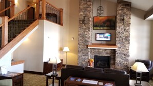 A spacious living area with a fireplace, a television mounted on a stone wall, a staircase with wooden railings, and a cozy seating arrangement.