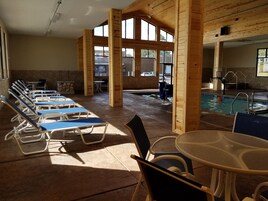 Amplia zona de piscina interior con sillones reclinables iluminados por el sol y una mesa de billar.