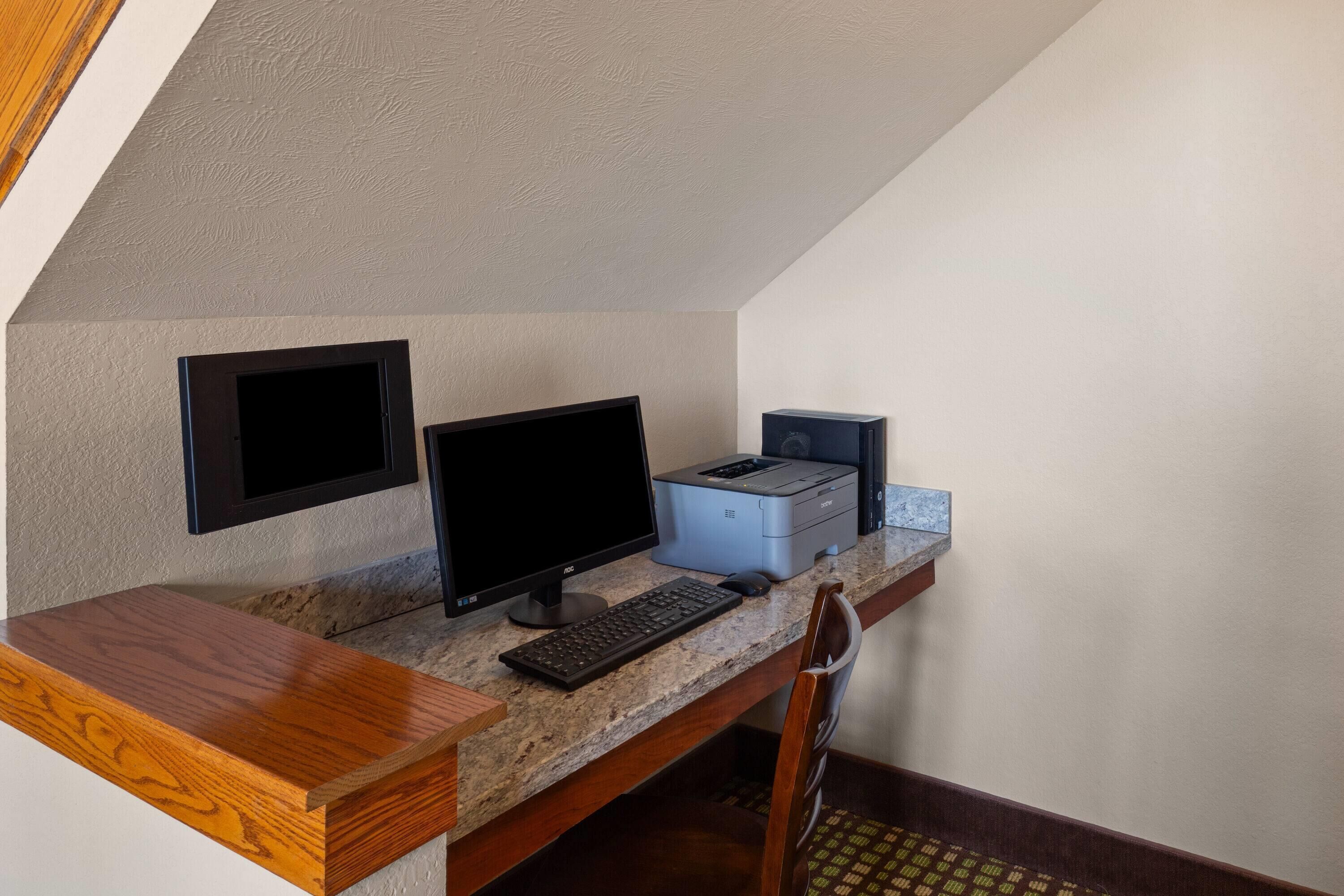 A computer desk with a monitor, keyboard, and printer in a room with a sloped ceiling and a chair.