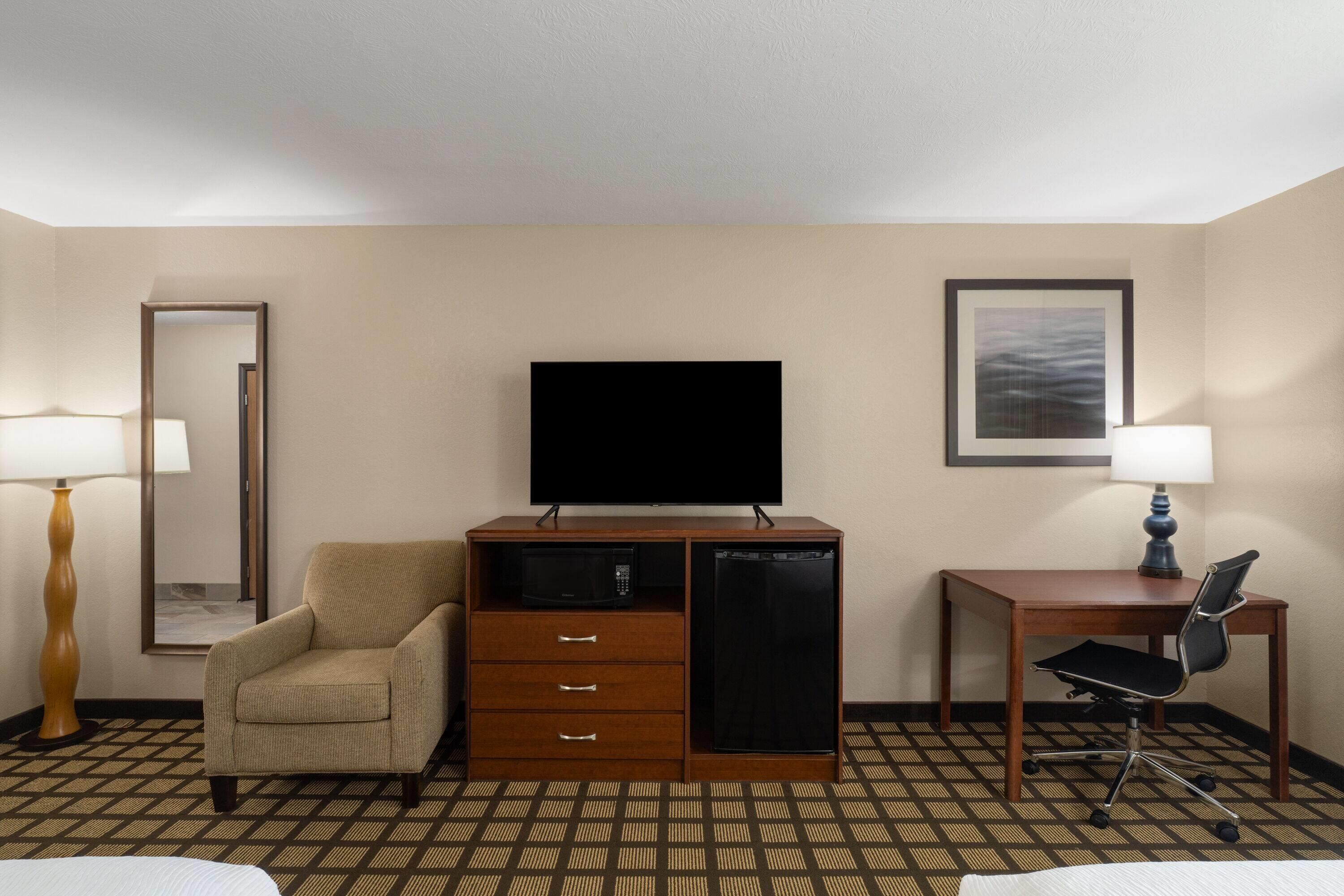 A hotel room with a bed, a desk with a chair, a television, a minibar, and a mirror.