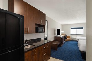 A compact hotel room with a kitchenette, a bed, a desk, and a window.