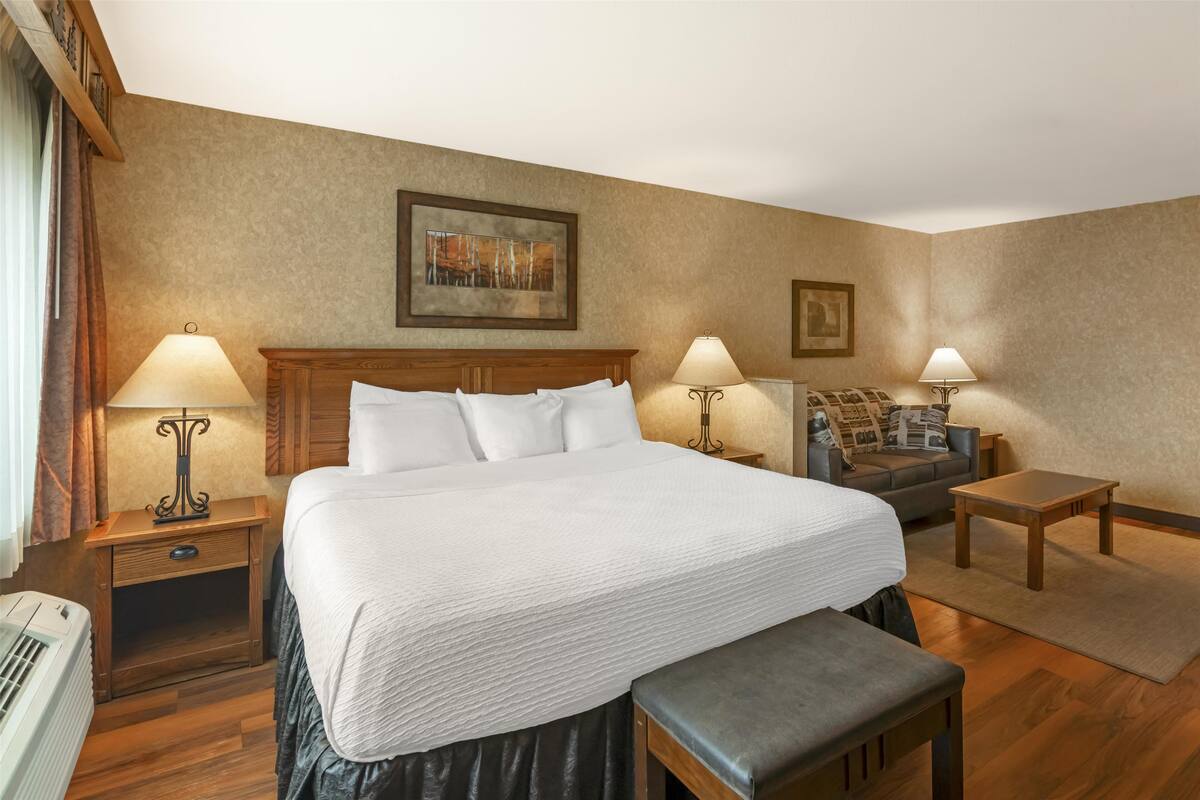 A hotel room with a large bed, two bedside tables with lamps, a sofa, a bench, and a rug.
