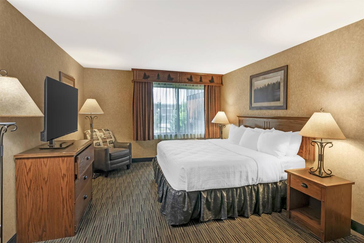 A hotel room with a large bed, two bedside tables with lamps, a desk with a television, a chair, and a window with curtains.