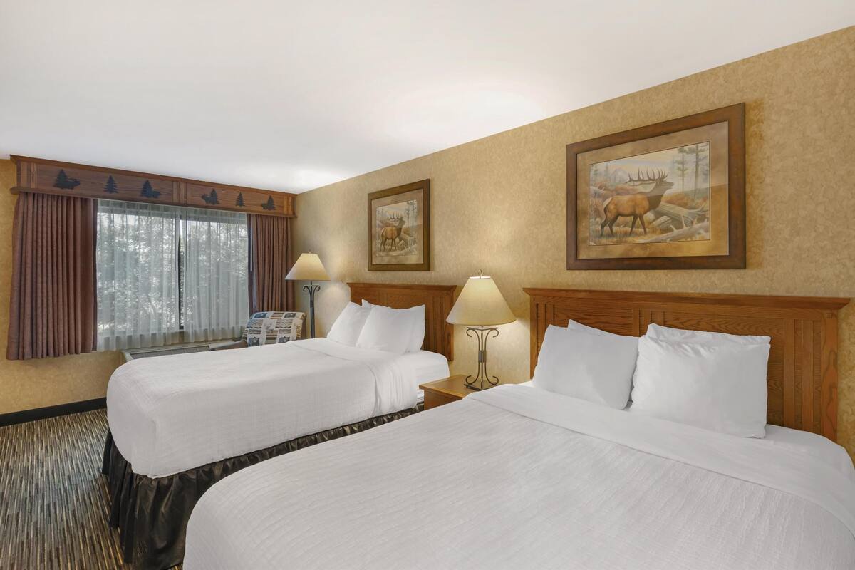 A hotel room with two beds, a large window with curtains, a wooden headboard, and two wall-mounted lamps.