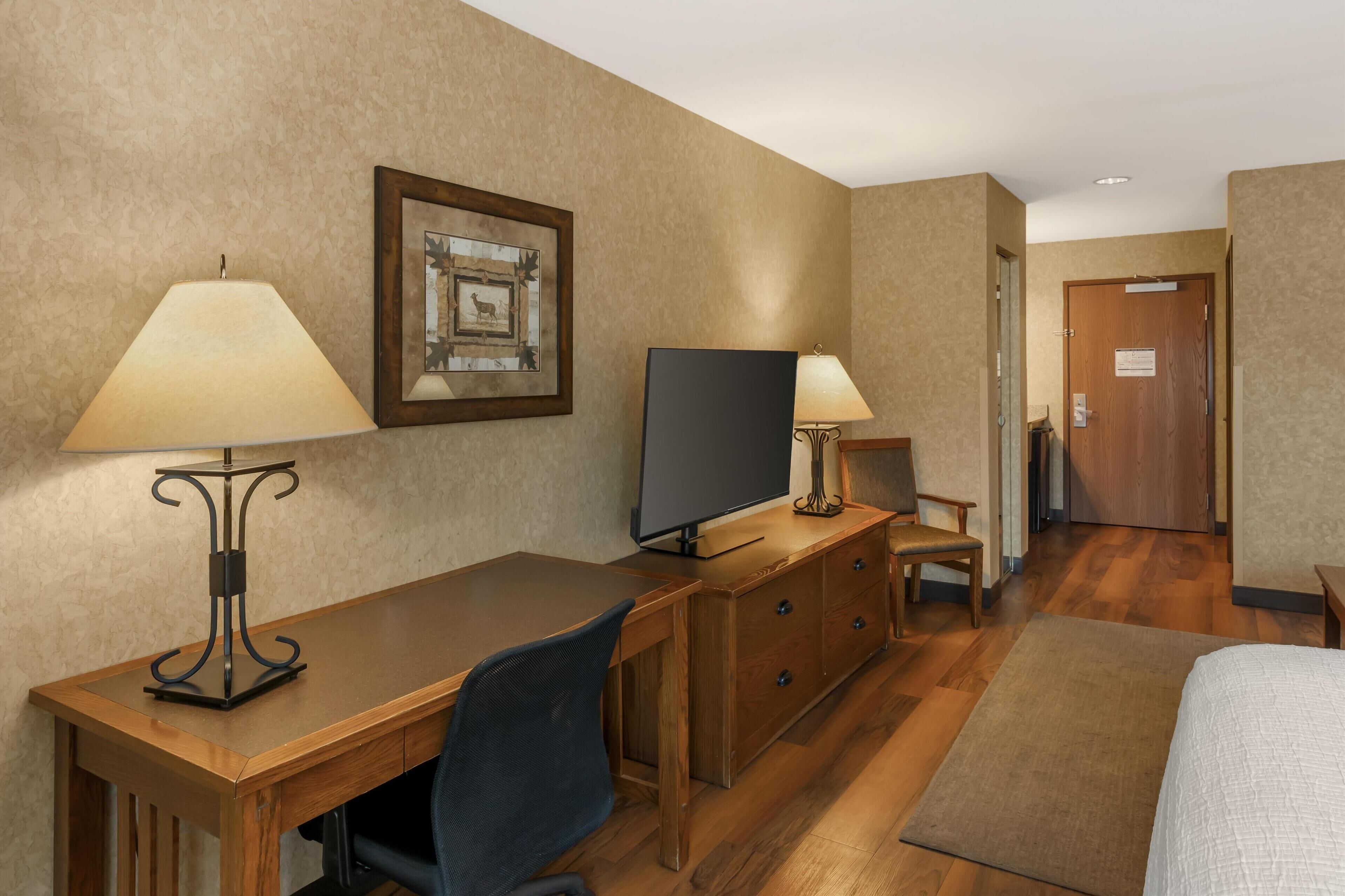 Una habitación de hotel con un escritorio de madera, dos lámparas, un televisor, una silla, una cama y una puerta.