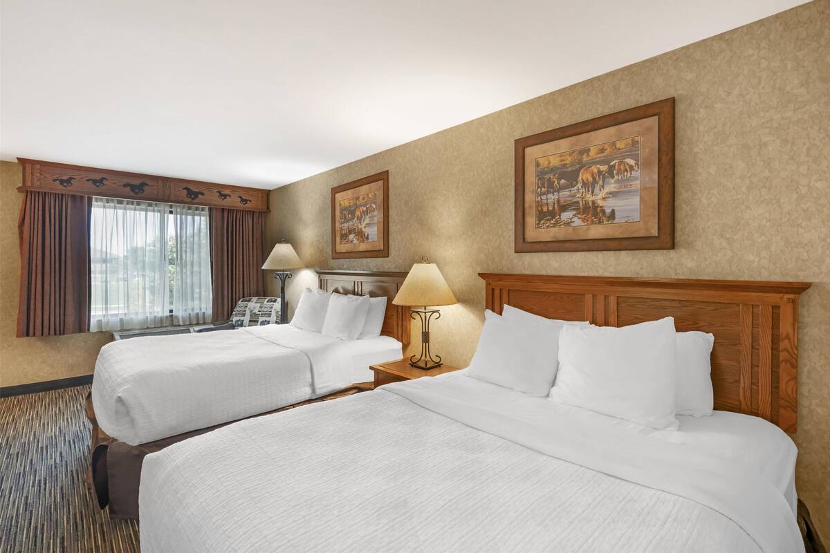 A hotel room with two beds, a wooden headboard, a window with curtains, a lamp, and framed pictures on the wall.