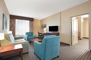 Una habitación de hotel con sofá, sillón, televisor, escritorio y una alfombra estampada.