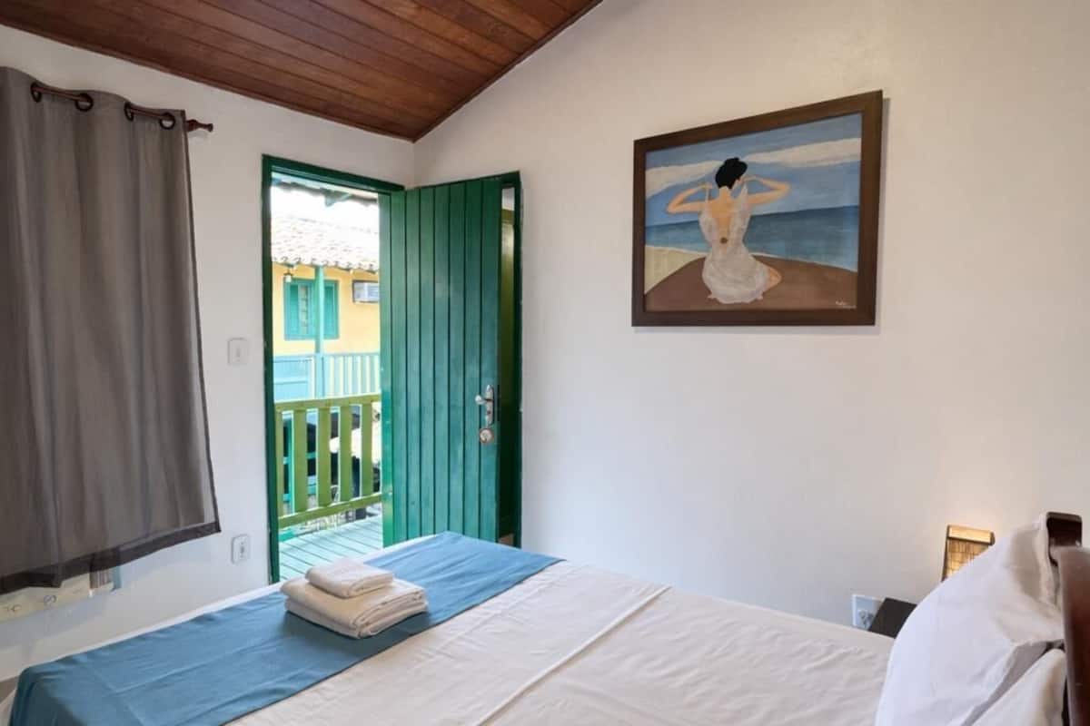 A bedroom with a bed, a painting, a window with curtains, and a wooden ceiling.
