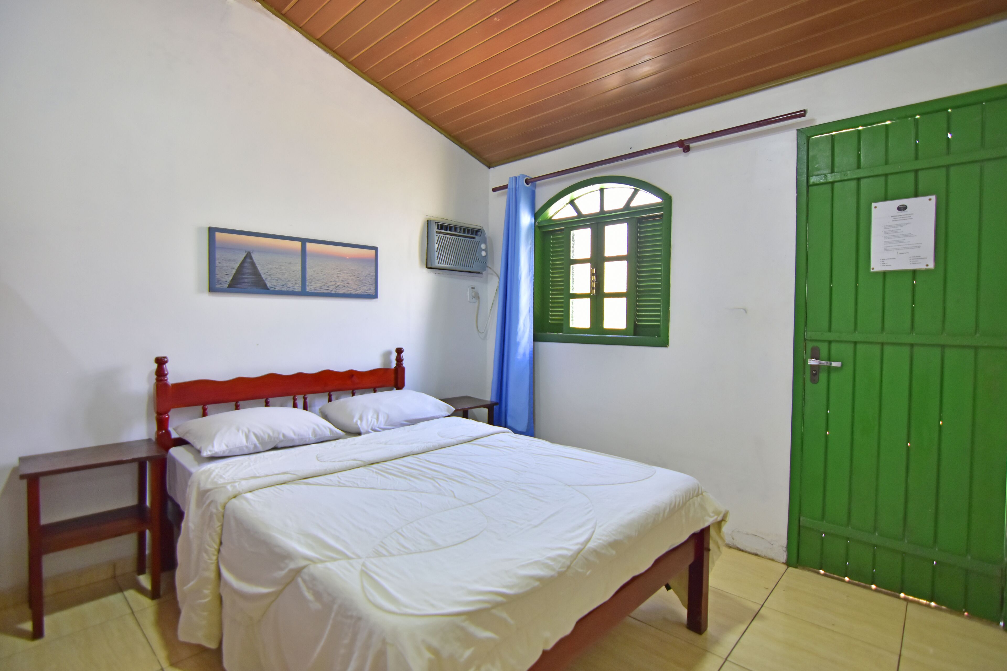Un dormitorio con cama de madera, una mesita de noche, una puerta verde, una ventana con contraventanas verdes y un aire acondicionado de pared.