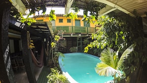 Un área de piscina con cubierta de madera, rodeada de plantas en macetas y una hamaca.