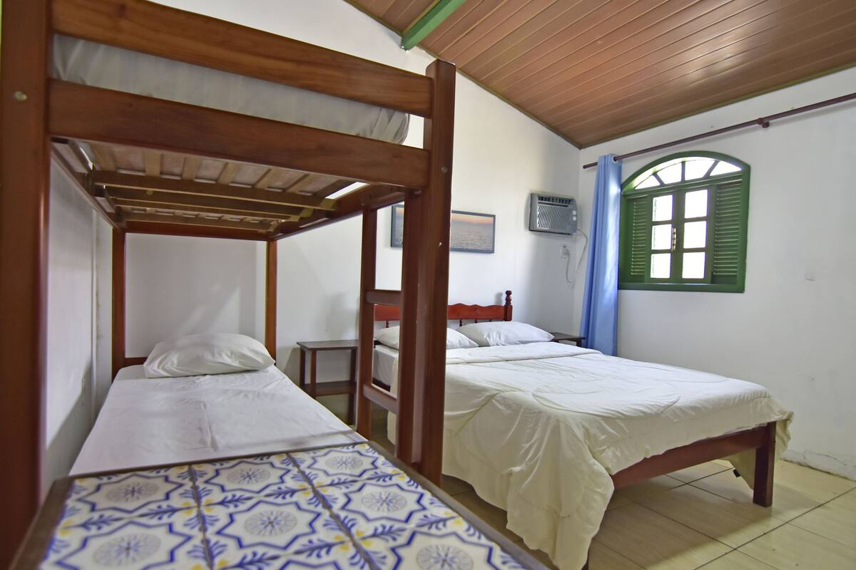 A room with two wooden bunk beds, a small table, a window with green shutters, and a wall-mounted air conditioner.