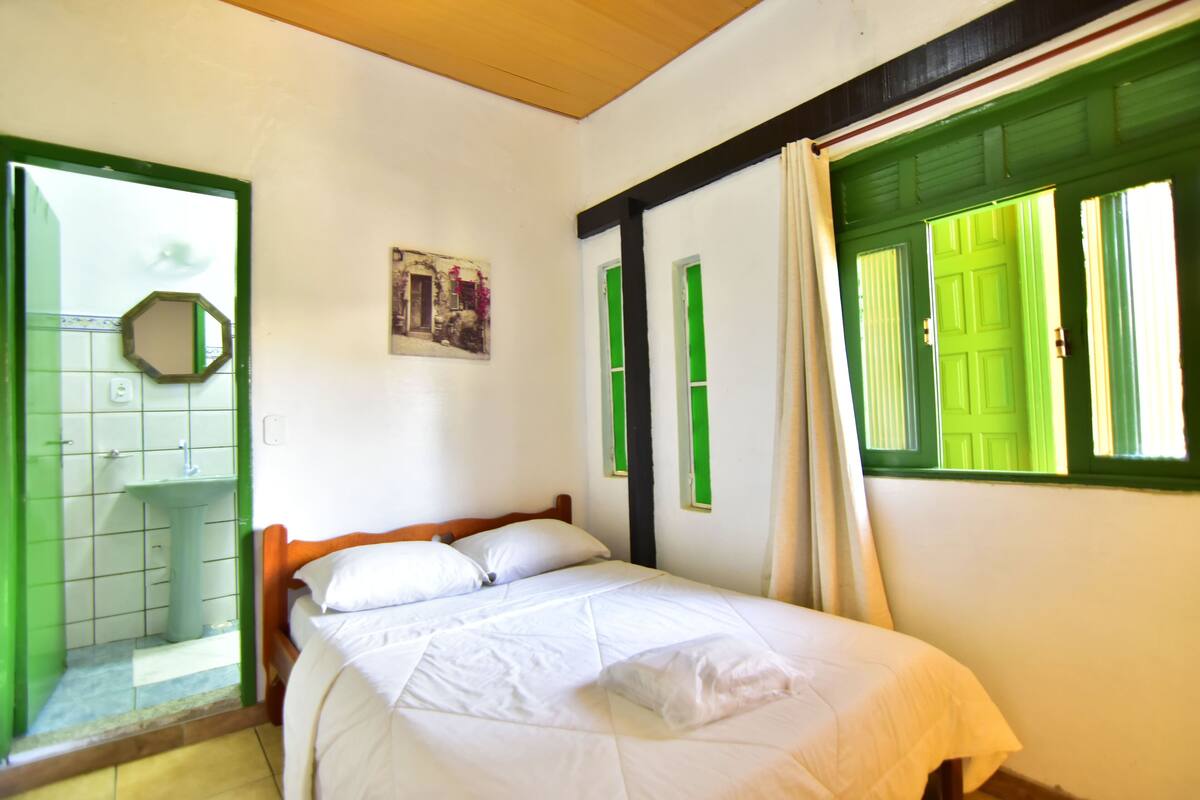 A room with a bed, a green door, a white sink, and a window with green shutters.