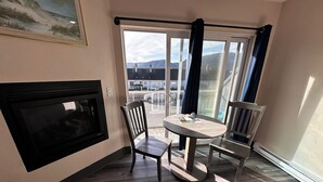 A room with a fireplace, a round table with chairs, a large window with a view of buildings and mountains, and a ceiling-mounted TV.
