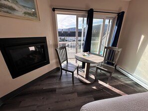 A room with a fireplace, a round table with chairs, a large window with a view of buildings and mountains, and a ceiling-mounted TV.