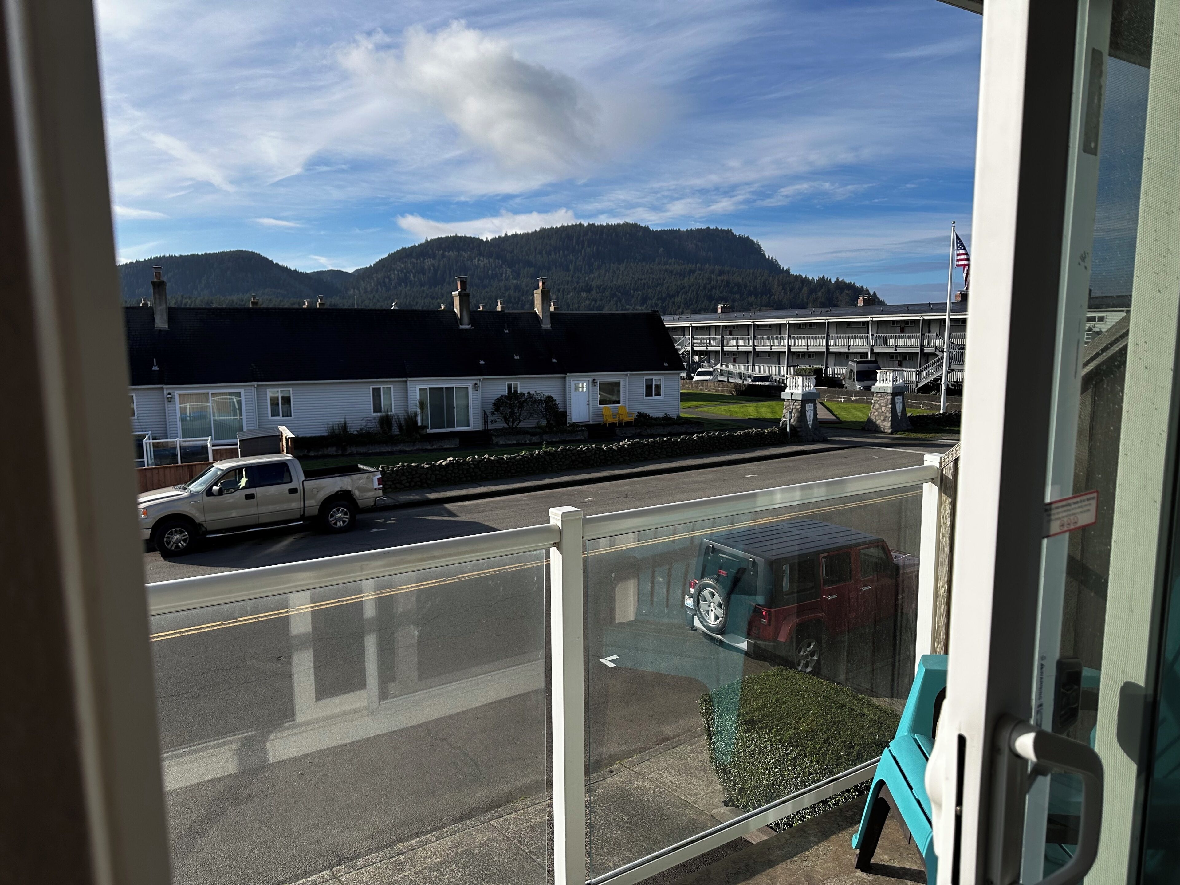 A view from a window overlooking a street with a white truck parked on the side, a building with a black roof, and a mountain in the background.