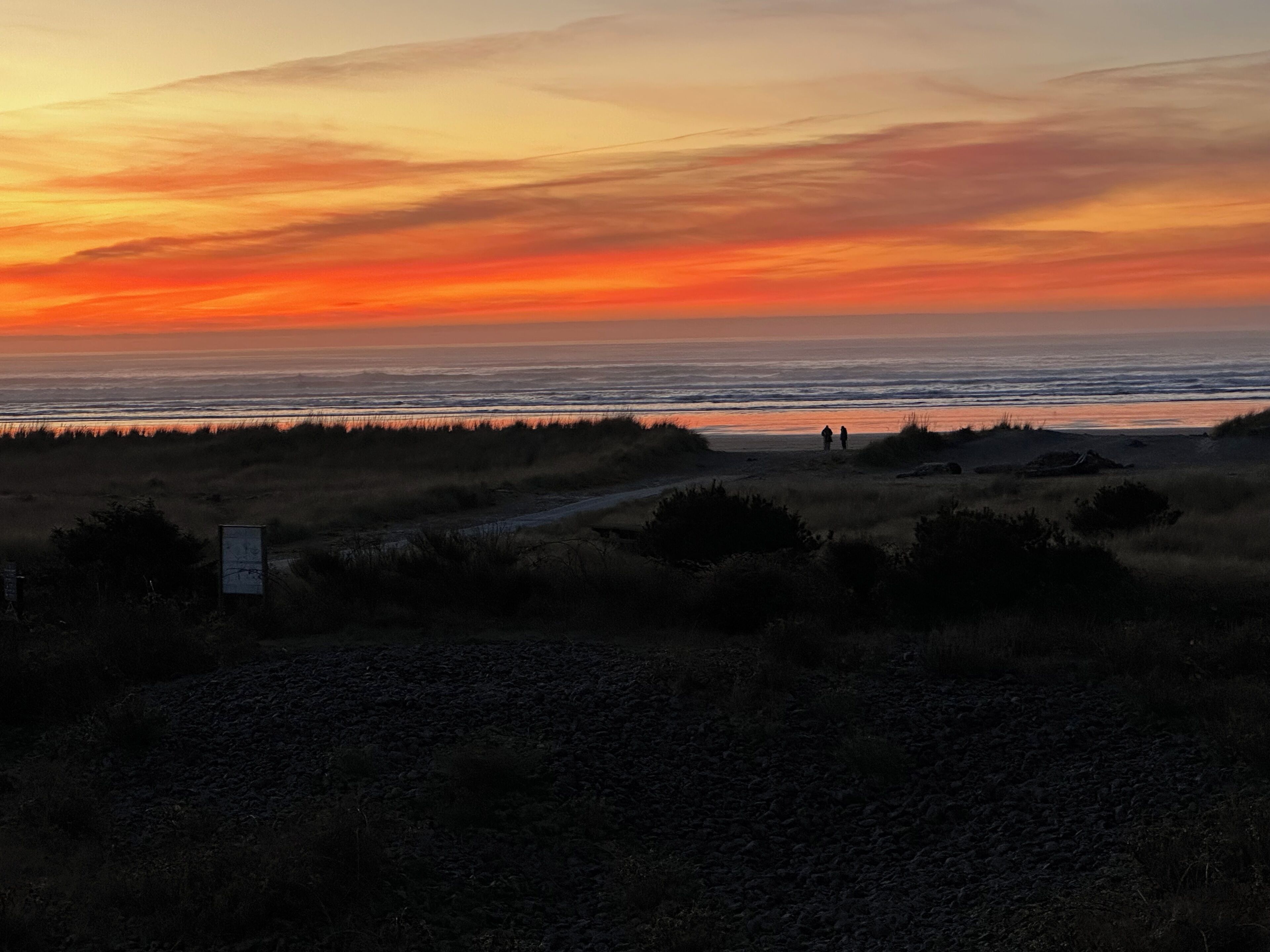 A beach scene with a sunset, two people walking, and a sign in the foreground.
