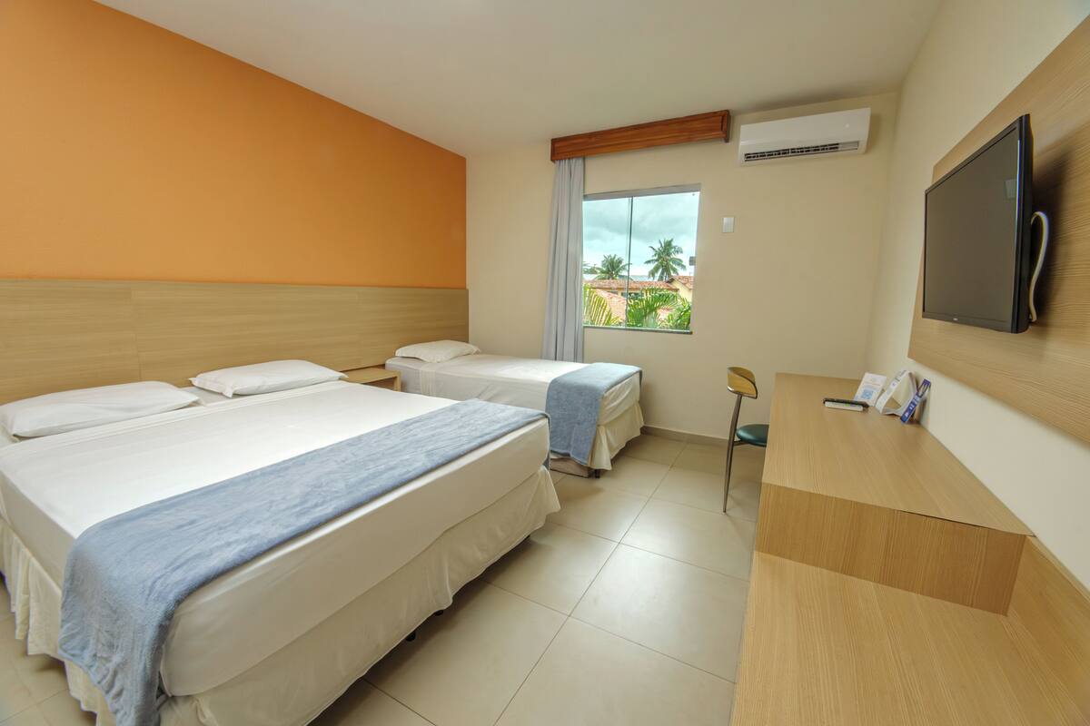 Habitación de hotel con dos camas, un escritorio, un televisor y ventana con vista a palmeras.
